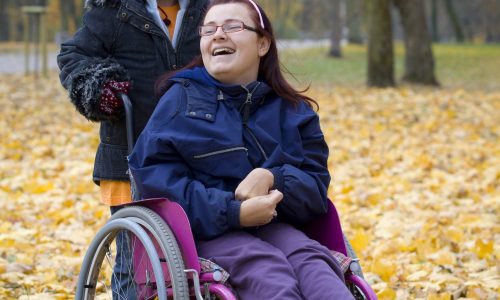Photo of two girls in a park. One girl is pushing the other girl who is in a wheelchair.