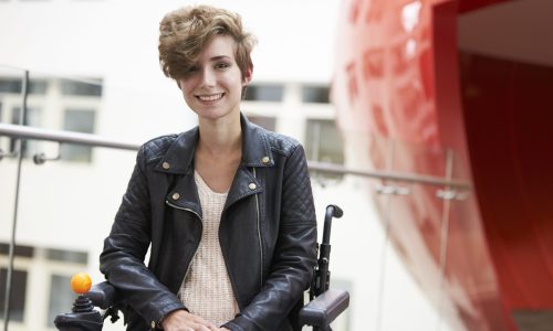 Photo of a woman in a wheelchair smiling. She has short brown hair and is wearing a leather jacket.
