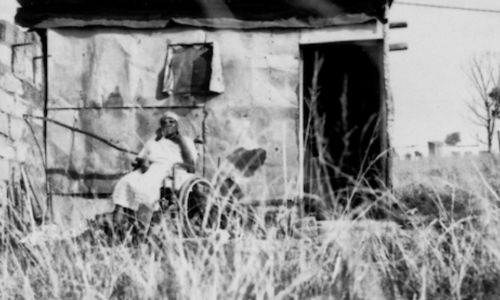 Photo of a woman in a wheelchair outside a disintegrating house.