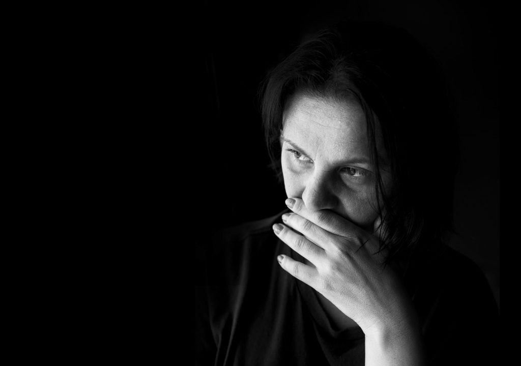 Greyscale photo of a woman with her hand to her mouth sitting in the dark.
