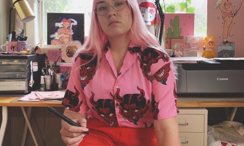 Photo of Ailie Banks, a young woman with pink hair, glasses, wearing a bright pink patterned top and bright red trousers sitting at her computer desk.