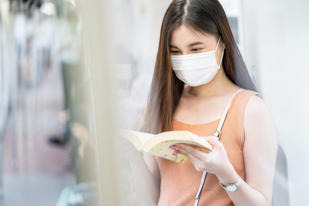 Young Asian woman passenger wearing surgical mask and reading the book in subway train when traveling in big city at Covid19 outbreak, Infection and Pandemic concept