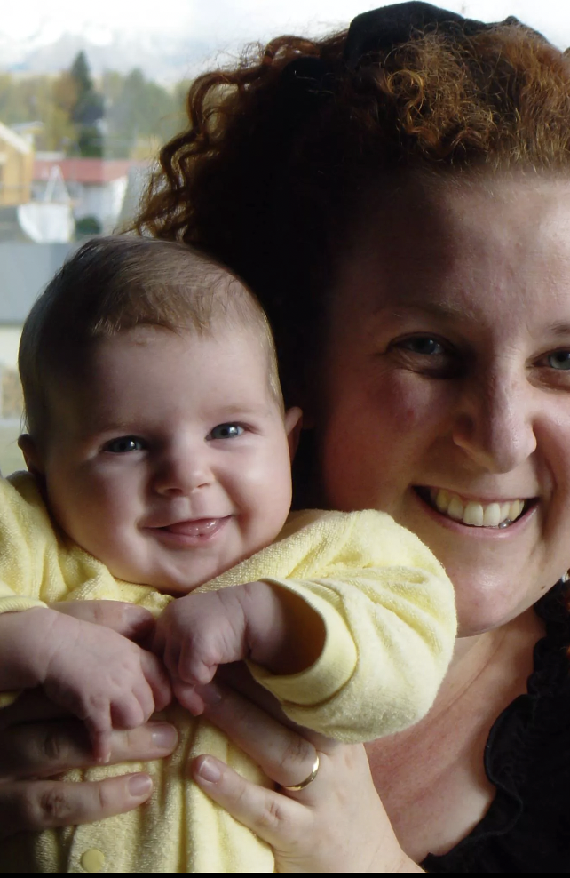 Issy (author) as a baby being held up by their mum. Issy is wearing a yellow grow suit and smiling with their tongue half out. Their mum is wearing a black top and has brown curly hair. She is smiling as well. 