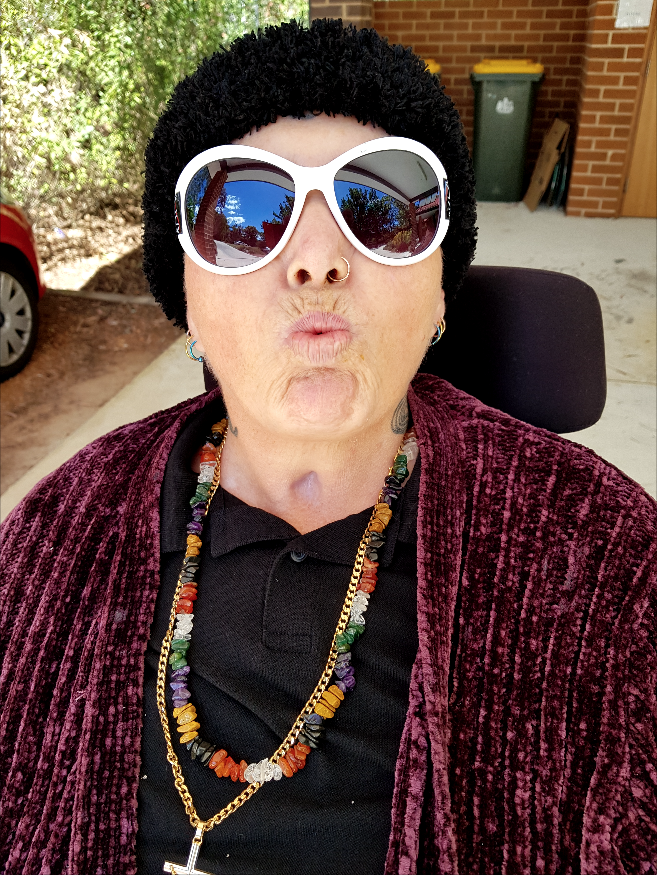 A photo of Julie, a fair-skinned woman wearing white sunglasses, a nose ring, a colourful necklace and a purple cardigan. She is pouting her lips and sitting in a wheelchair. 