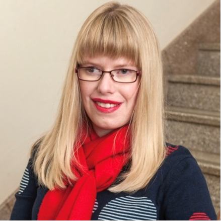 Jane Britt (image description: a woman smiling with blonde hair and glasses. Jane has straight hair and a fringe and is wearing a blue jumper and a red scarf.