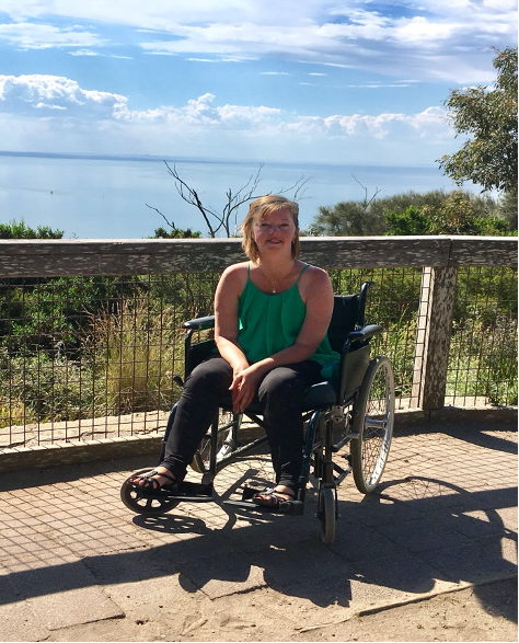 Karen Fankhauser: (image description) a woman sitting in a wheelchair in front of a fence, with the ocean in the background. Karen is wearing a green top, black jeans has light brown hair and is smiling.