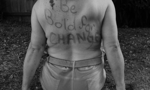 Photo of Tammy Milne, sitting on grass, facing away from the camera with the words "Be Bold for CHANGE" written in texter on her back.