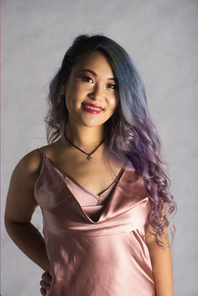 a person with long blue and purple hair who is smiling. Akii is wearing a pink dress. 