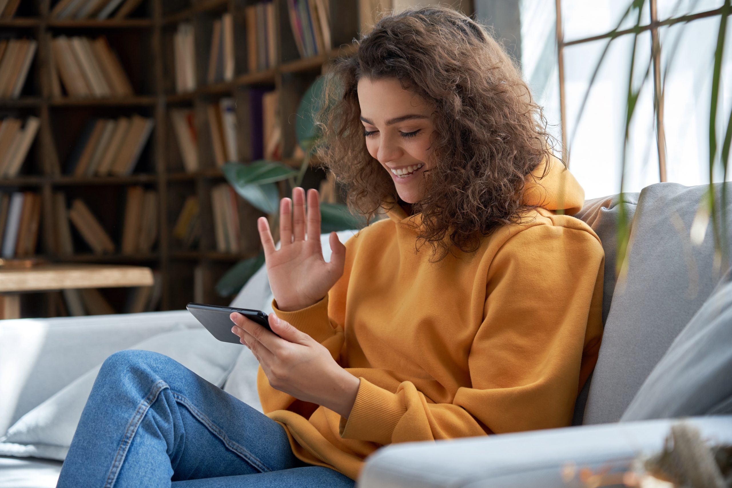 photo of a young woman using a smart phone while sitting on a couch. The young woman has dark curly hair and Is wearing a yellow jumper and denim blue jeans. She is smiling and waving at the phone.