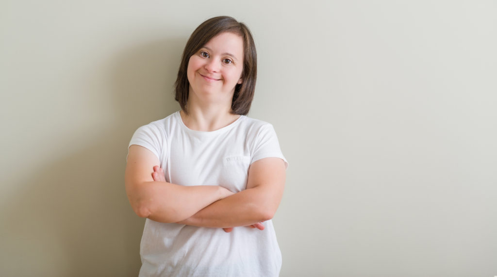 Woman with Down Syndrome wearing a white top standing against a wall smiling. 