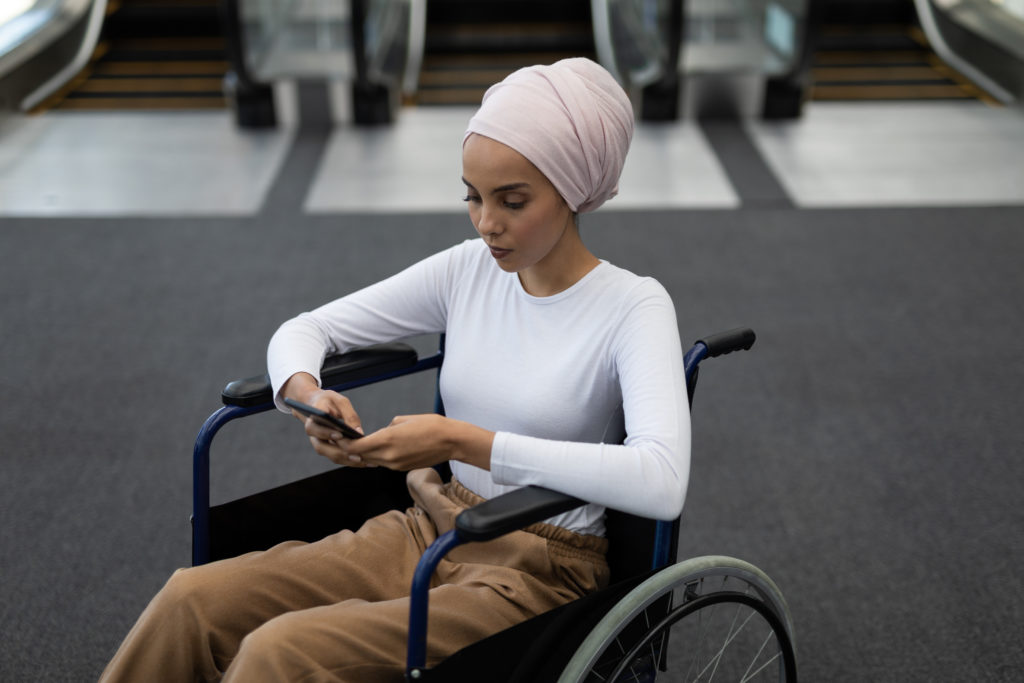 A woman sitting in a wheelchair, wearing a pink head scarf and white top, using her phone.