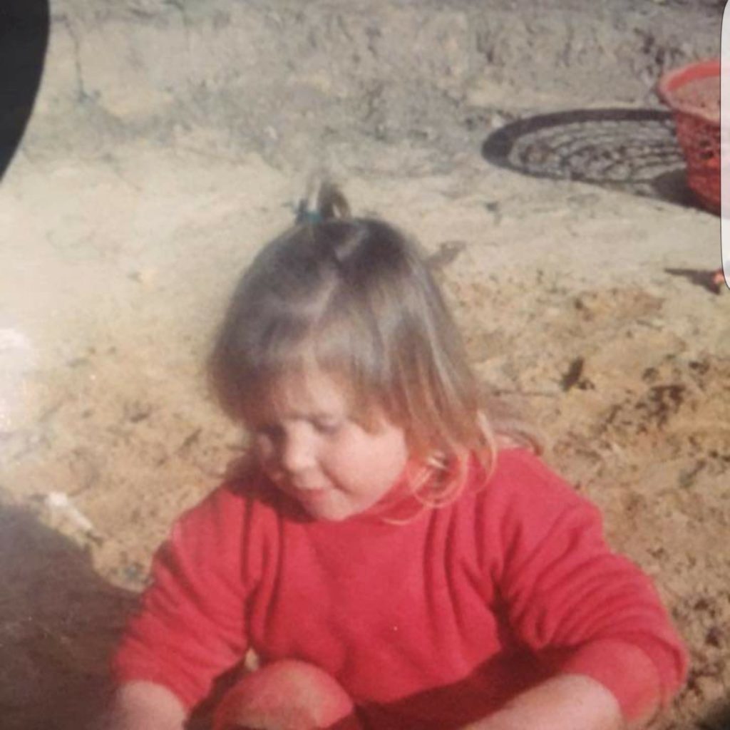A young Indigenous girl with light brown skin and dark blonde hair, wearing a red jumper, is crouching down to play at the beach. She is looking at what she holds and behind her is a red plastic washing basket sitting on the sand.