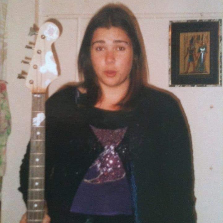 A teenaged Indigenous girl, with light brown skin and dark brown hair stands holding a bass guitar awkwardly. She is wearing a sparkly purple top under a fluffy black jumper.
