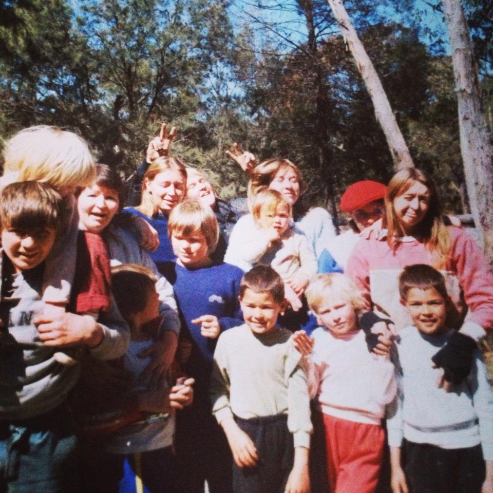 A large group of people stand close together for a family photo with a mix of adults and children. They all are wearing various 1980s style tracksuits. Behind them is thick bushland.