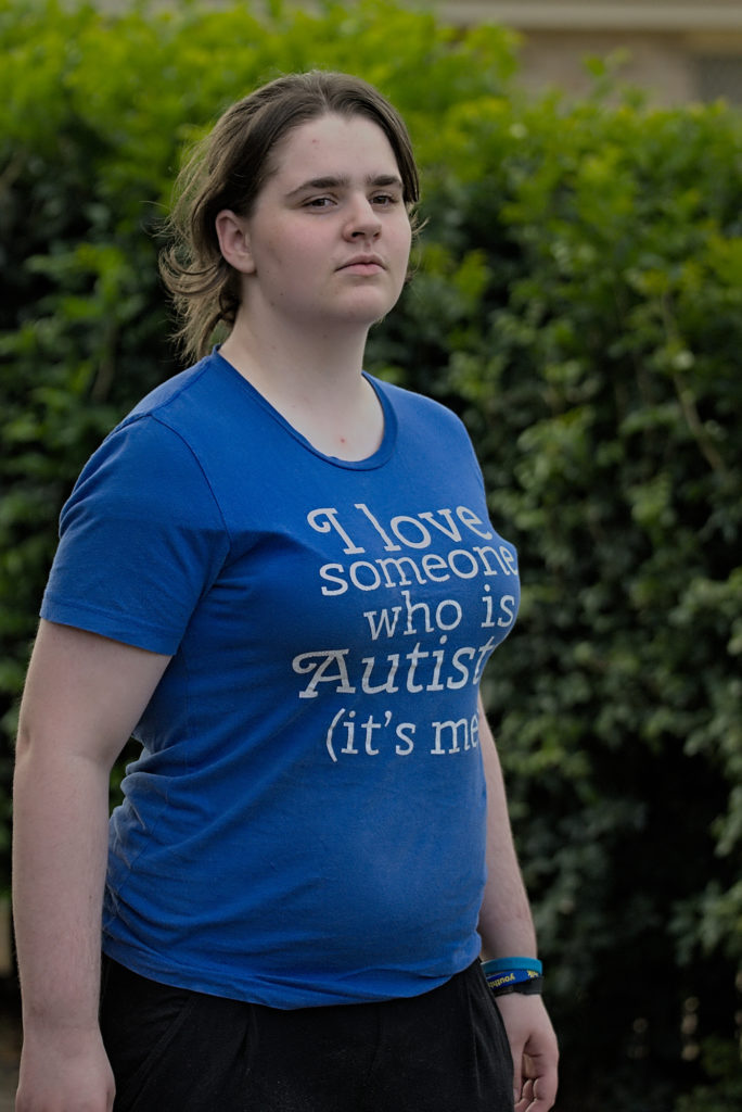 Photo of Maria who has dark brown hair tied back in a ponytail and is wearing a blue shirt that reads I love someone who is autistic (it's me).