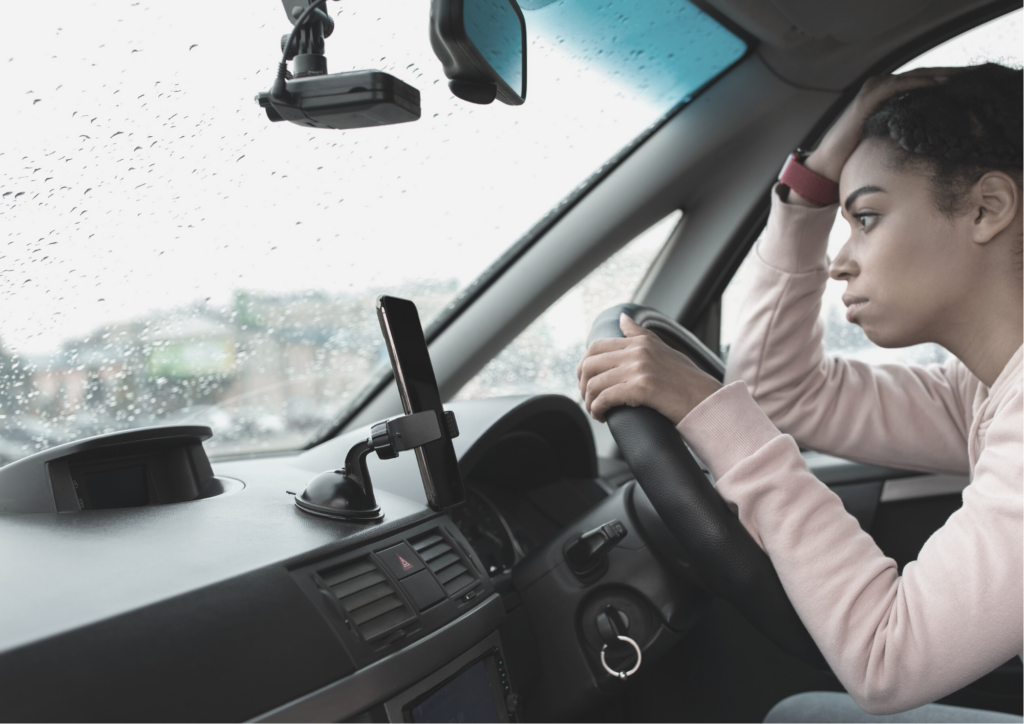 A photo of a women driving a car, but leaning on the steering wheel stressed.