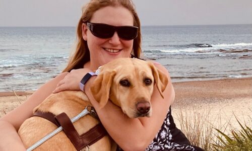 Photo of Katie who has long blonde hair and is wearing dark sunglasses. She is hugging her dog called sadie who is wearing a mobility support harness.