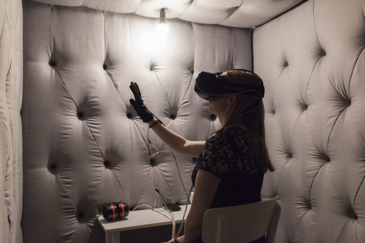 Image description: In a grey sound-proofed room with dim lighting, a woman sits at a desk. She is wearing a virtual reality headset and holding up her hand, which is wearing a custom-built glove. On the desk are different cables and speakers.