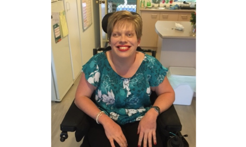 A photo of Melanie Schlaeger with short hair, red lipstick and a flowery blue top sitting in a wheelchair.