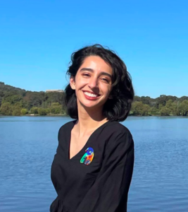 A young woman with short black hair and brown skin wearing a black V neck with full sleeves and a colourful brooch of a bird. She is smiling and the background top is blue sky and bottom is blue water.