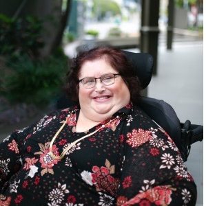 Photo of Karin Swift who has short black/ red hair and is wearing glasses with a black red floral top on. She is sitting in her wheelchair which trees in the background.