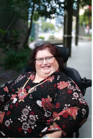 Photo of Karin Swift who has short black/ red hair and is wearing glasses with a black red floral top on. She is sitting in her wheelchair which trees in the background.