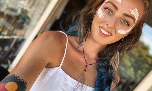 Lily, a proud Wiradjuri woman, is looking into the camera, smiling. She has long brown curly hair with blue tips. She is wearing white ochre on her face, along with emu feather earrings and an emu feather necklace. On her right arm is a tattoo of the Aboriginal flag.