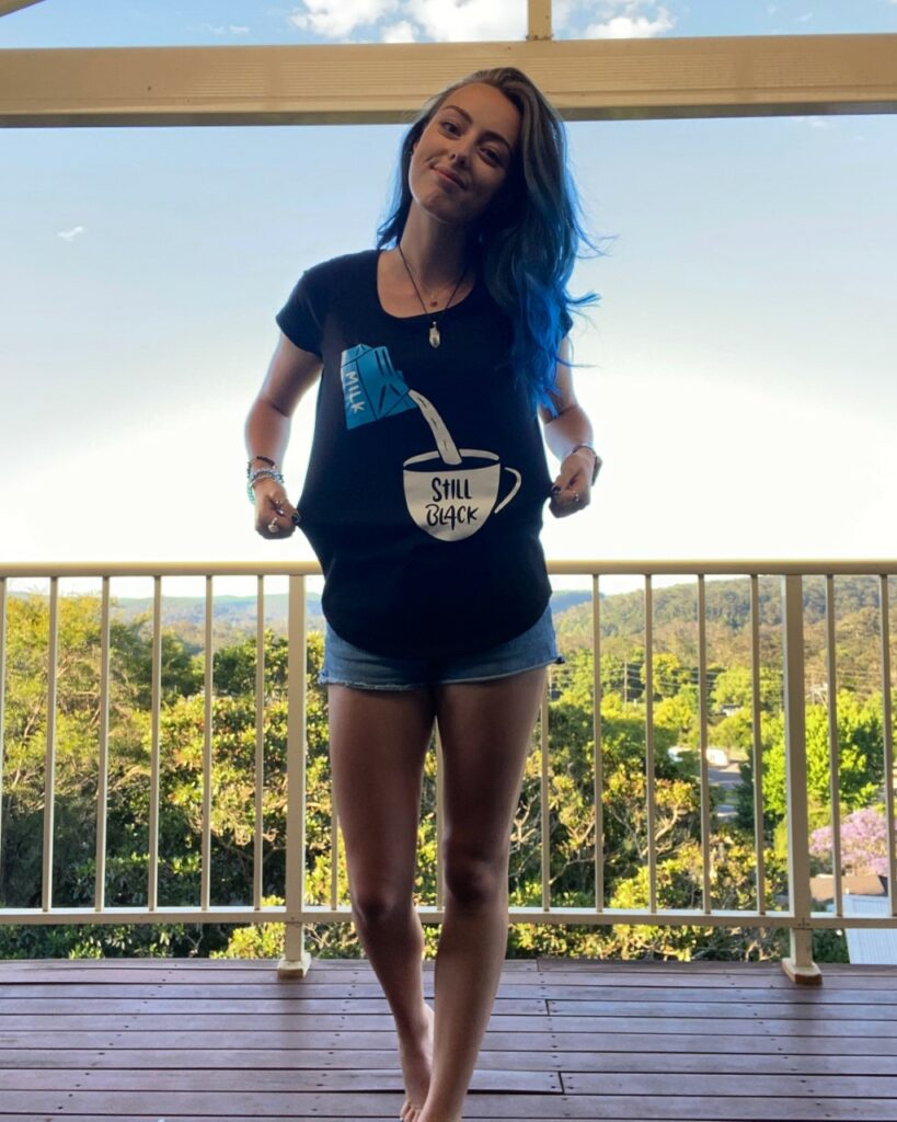 Lily, a proud Wiradjuri woman, is standing on a balcony. She has long brown and blue hair. She is holding out her t-shirt to draw attention to it – on it is an illustration of milk being poured into a cup of coffee that reads “Still Black” on the mug.