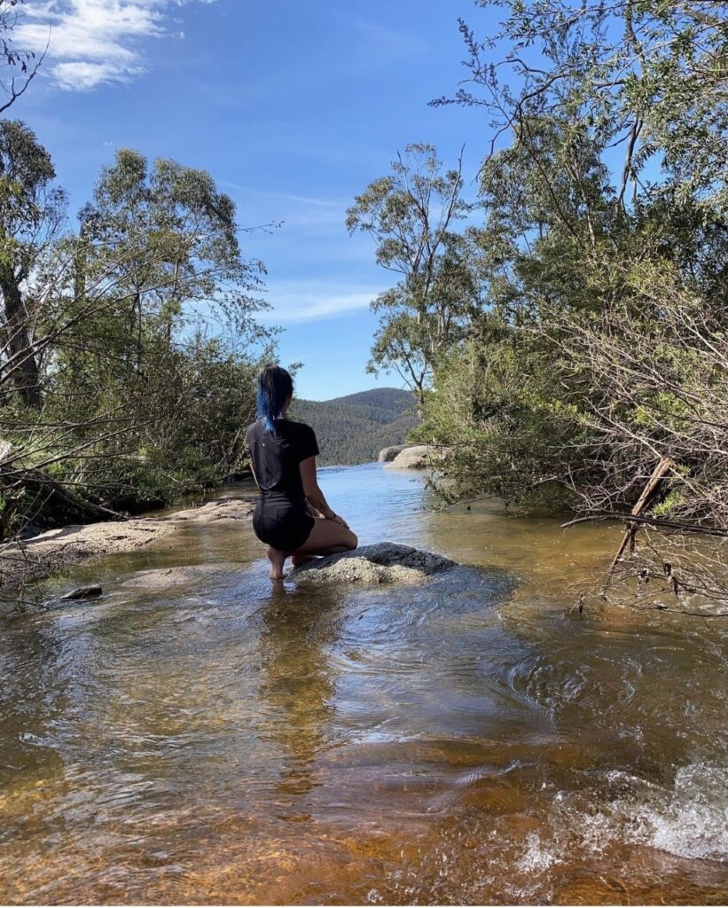 Lily, a proud Wiradjuri woman, is kneeling on rocks within a flowing creek. She is wearing all black and has her back to the camera. On either side of the creek are trees hanging overhead, and in the distance behind Lily you can see mountains. 