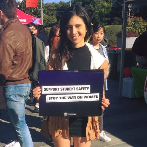 Photo of 23 year old Heidi at University of Sydney O’Week holding a purple sign with highlighted text reading: ‘Support Student Safety. Stop The War On Women.’ Heidi is wearing a black top and skirt and a brown coat. She has long dark brown hair and is smiling.