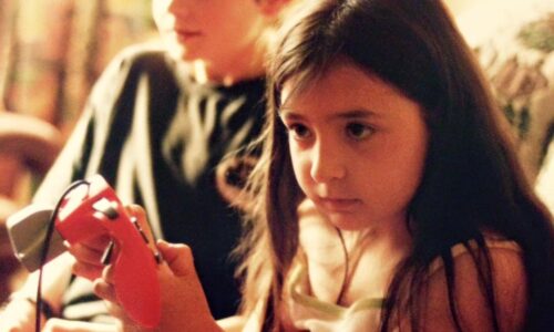 Photo of seven year old Heidi sitting on a couch with her cousin Alec, a young boy. Both Heidi and Alec are looking to the left immersed in what they are doing. They are both holding Nintendo controllers. Heidi has long dark brown hair and is wearing a white and pink dress. Her cousin in the background is wearing a black top and has short light brown hair.