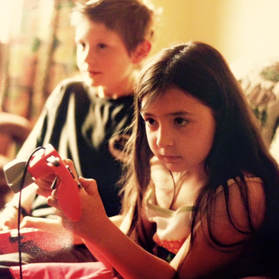 Photo of seven year old Heidi sitting on a couch with her cousin Alec, a young boy. Both Heidi and Alec are looking to the left immersed in what they are doing. They are both holding Nintendo controllers. Heidi has long dark brown hair and is wearing a white and pink dress. Her cousin in the background is wearing a black top and has short light brown hair.