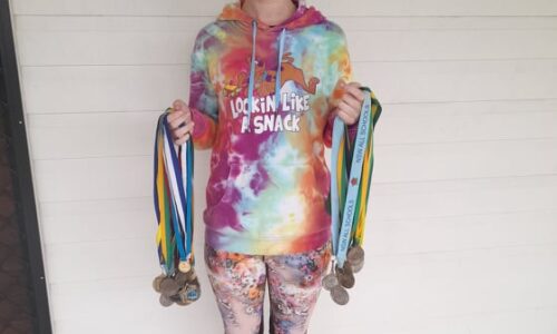 • A photo of Steph, a white woman with short brown hair, holding dozens of sporting medals. Steph is wearing a tie-dye hoodie and colourful leggings.
