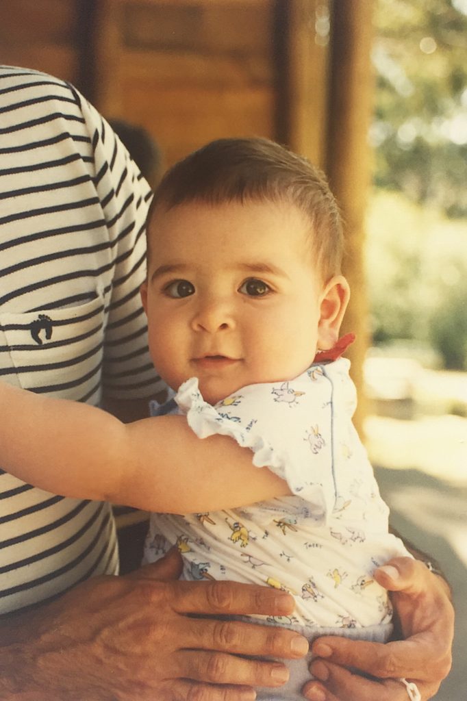 Heidi as a one year old baby being held by her father. She is wearing a white top with cartoons of birds on it and is smiling at the camera. She is has short brown hair and brown eyes.