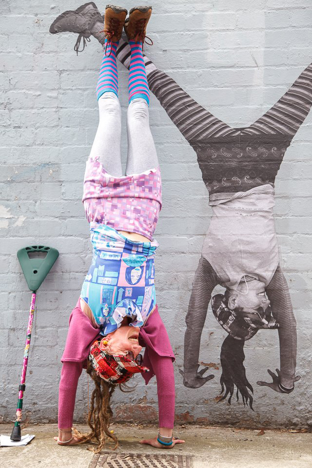 A photo of Larissa smiling and doing a handstand against a brick wall. She is wearing colourful pink and blue clothing, and has her walking stick leaning on the wall next to her. Painted on the wall is an artwork of Larissa doing a handstand! 
Image credit: Larissa MacFarlane