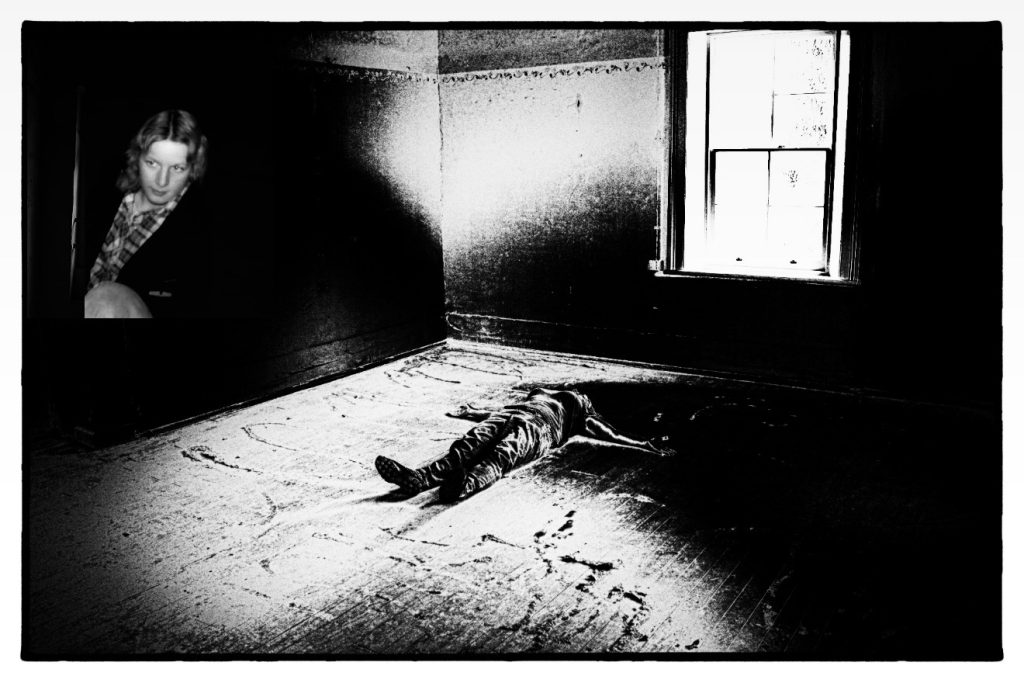 A black and white super-exposed photo of a person lying on their back on the floor, arms spread out, in the middle of a cell at the Kamballa Special Unit at Parramatta. On the wall of the room is a photo of a young woman.