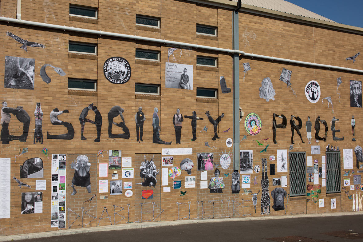 An image of the Disability Pride mural in Melbourne. It features many different pasteups on a brick wall, including the text 'Disability Pride' spelt out using images of different disabled people contorting to make the shape of letters.