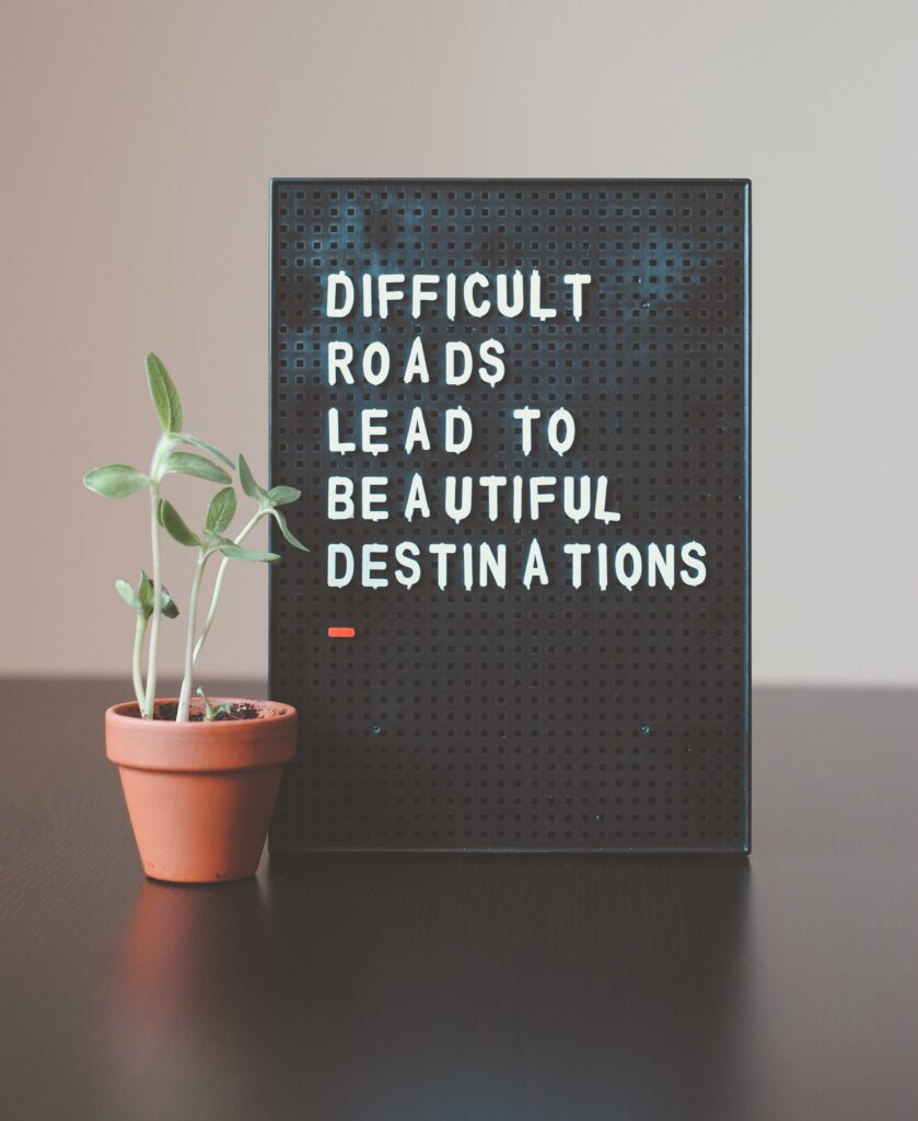 A black letter board stands on a table top, with white letters that read 'Difficult roads lead to beautiful destinations'. A growing plant is next to the letter board.