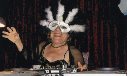 A small-statured woman wearing a black dress, see-through cardigan and white dress-up mask with feathers is smiling and laughing. She is sitting behind some DJ decks she is using at a masked-ball event.