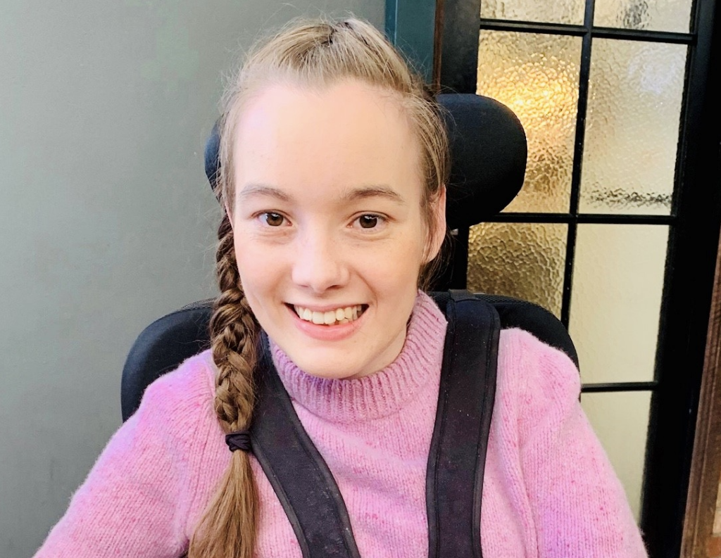 A photo of Claire, a white woman with fair skin, long brown hair that is braided to the side, wearing a pink jumper. Claire is sitting in a black power wheelchair, with a grey wall and frosted glass door behind her. 