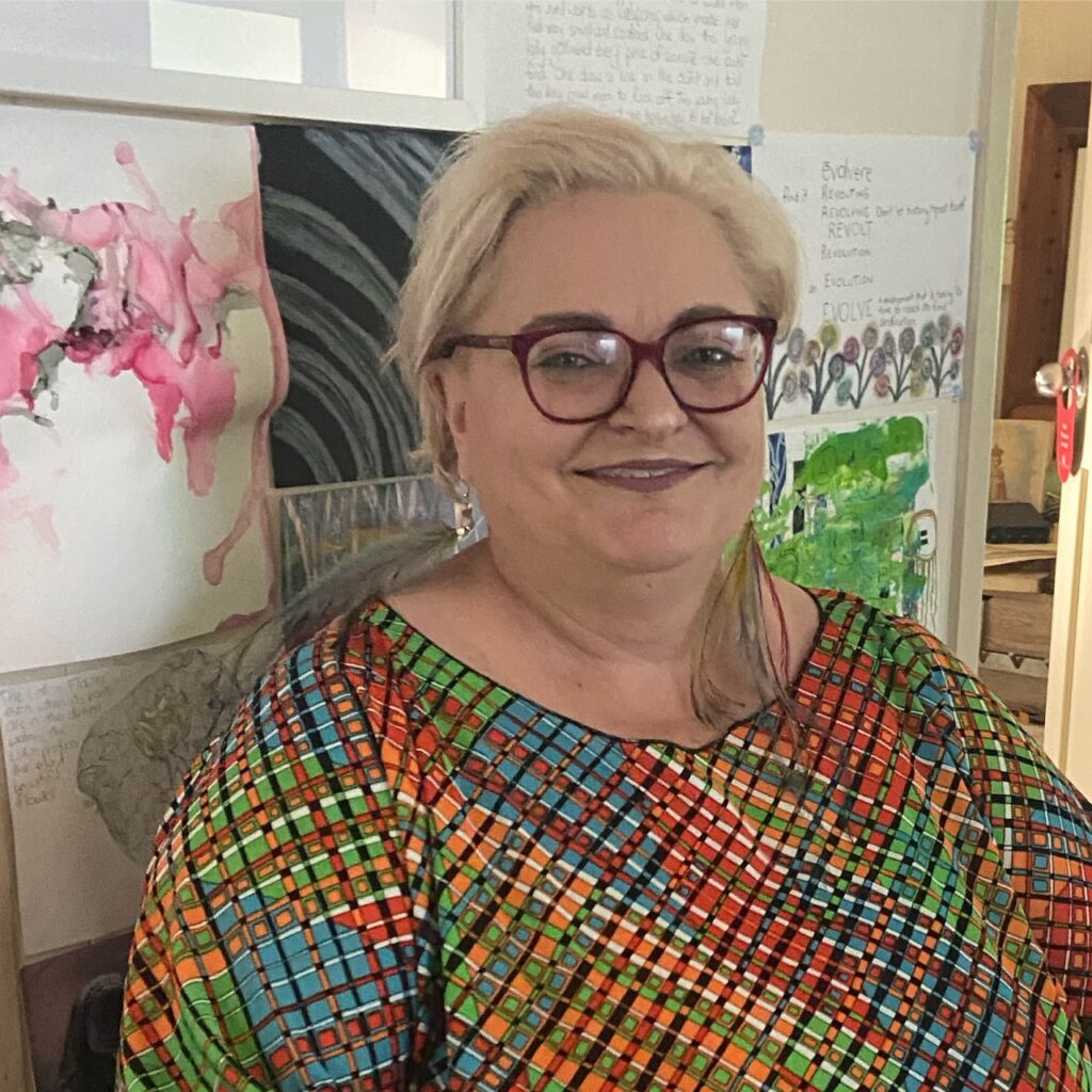 A middle aged white woman wearing pink framed glasses with short blonde hair. She is wearing a multicoloured geometrical Kaftan and standing in front of some artwork.