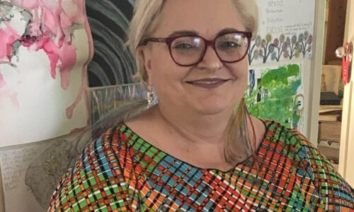 A middle aged white woman wearing pink framed glasses with short blonde hair. She is wearing a multicoloured geometrical Kaftan and standing in front of some artwork.