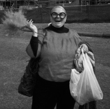 A middle aged white woman with blonde hair standing on an oval. She is wearing a top and pants with whacky glasses and holding a large feather and white plastic bag in either hand.