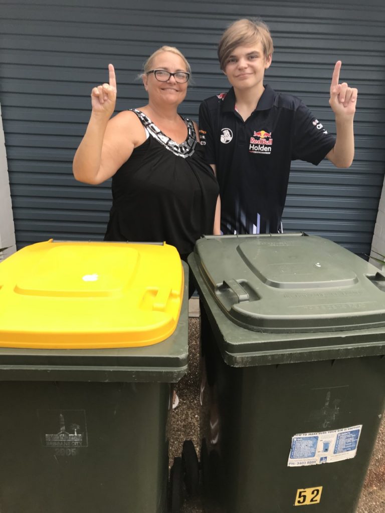 •	An image of Laura and her son standing in their driveway. They both have pale-skin and are wearing different black shirts, holding up a finger each to signify the number one. In front of them is a yellow recycling bin and green waste bin.