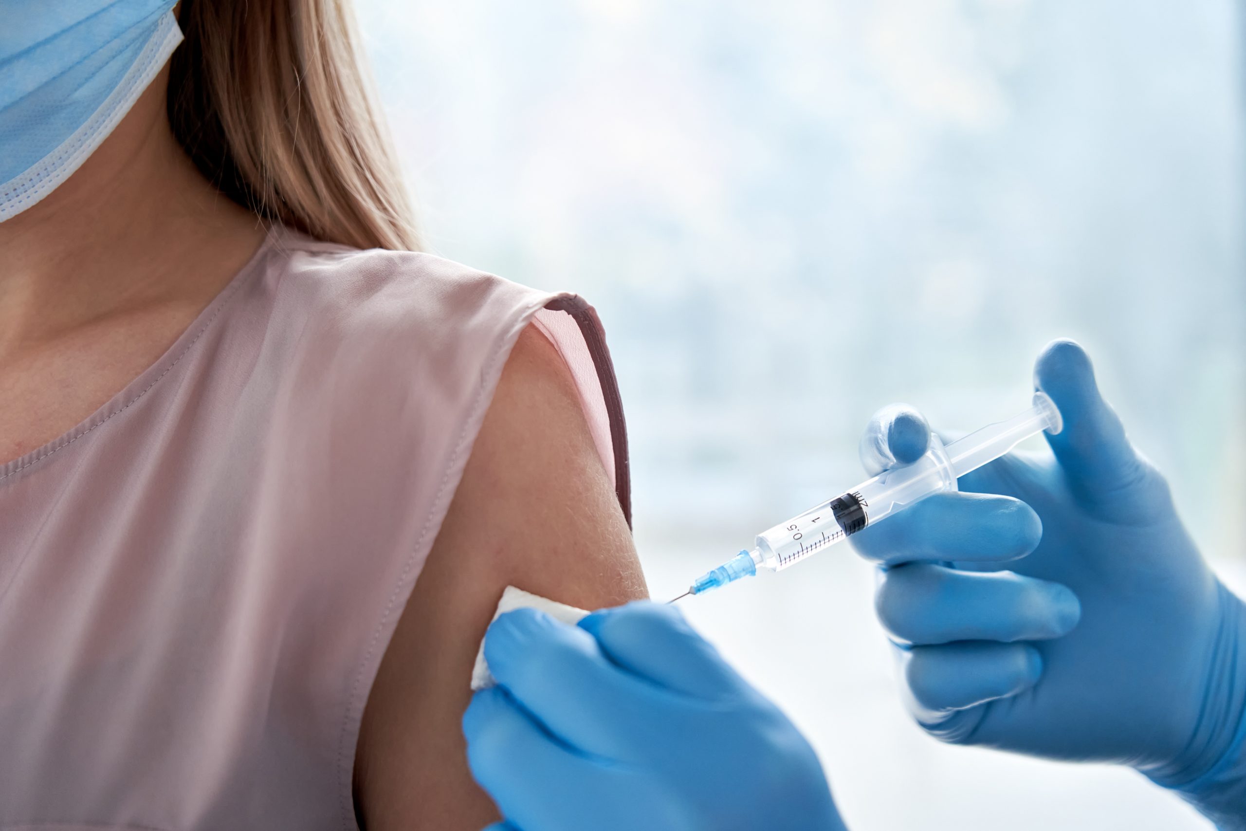 An image of a woman being injected with a COVID vaccine by a doctor wearing blue sterile gloves. Only the woman’s shoulder and neck are visible. She has white skin, blonde hair and is wearing a light pink, sleeveless blouse.