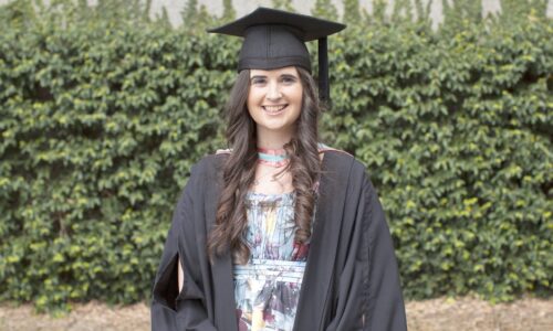 An image of Katherine smiling in front of greenery in graduation attire. Katherine has long brown hair that is curled, and is wearing a colourful floral dress, black graduation robe and black graduation cap.