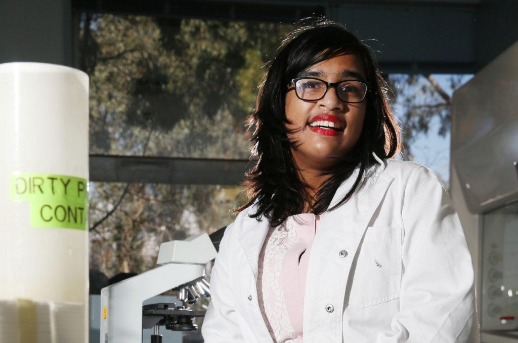 A photo of Jerusha smiling in a science lab. Jerusha has brown skin and black, shoulder-length hair. She is wearing glasses, red lipstick, a white lab coat and a pink dress. In the background microscopes and scientific equipment can be seen. 