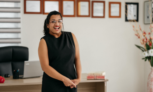 A photo of Jerusha standing and smiling at the camera. Jerusha has brown skin and black hair in a ponytail. She is wearing a black dress and glasses. In the background you can see an office desk, chair and laptop, as well as qualifications and degrees hanging on the wall.