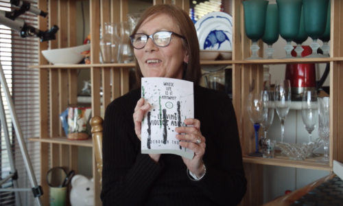 A photo of Deb Thomson holding a copy of her book 'Whose life is it anyway? Leaving a violent abuser' and smiling. Deb is a white woman with black glasses and shoulder length brown hair. She is wearing a black turtle-neck shirt. In the background is a cabinet with glasses, fine china and a telescope.