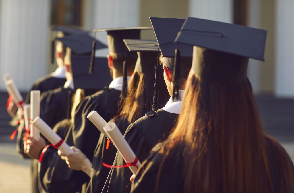 An image of a group of university graduates lining up, holding their degrees during a graduation ceremony. The photo is taken from the rear, so only the backs of students in black robes and graduation caps are visible.